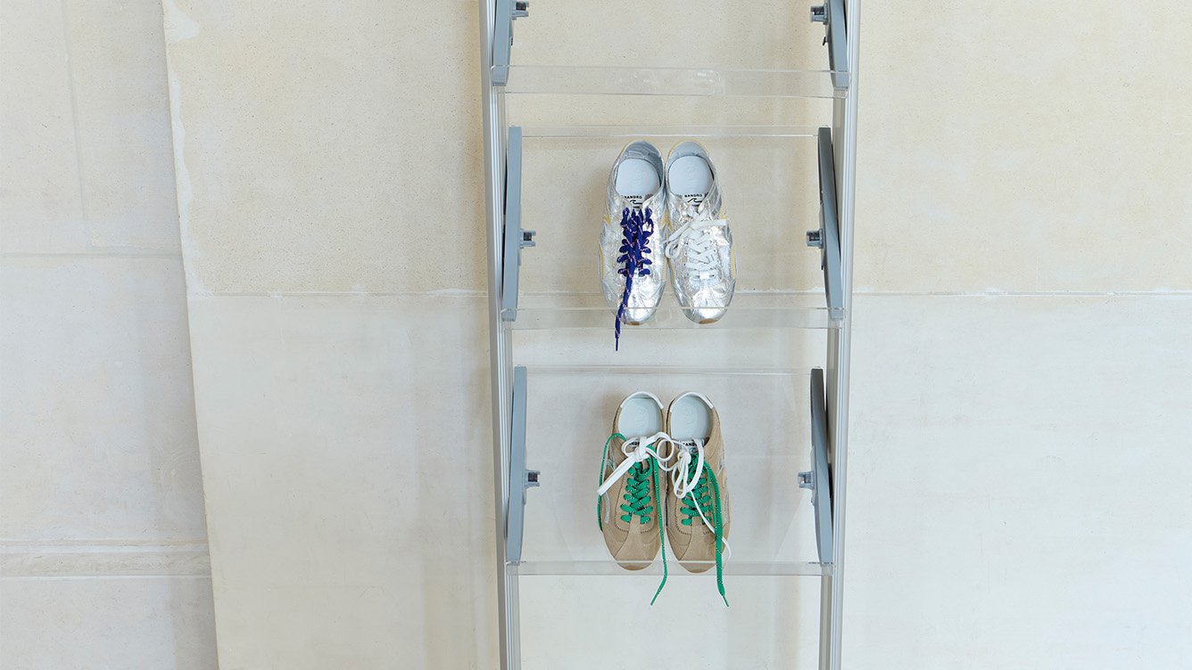 Two pairs of Sandro Light Wave sneakers displayed on transparent acrylic shelves: one silver pair with blue laces (top) and one beige suede pair with green laces (bottom), from the SS26 collection.